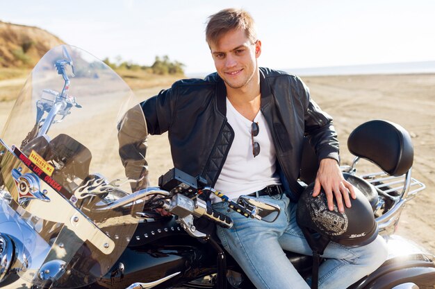 Ciérrese encima del retrato del motorista en el camino cerca de la playa. Hombre guapo sentado en moto.