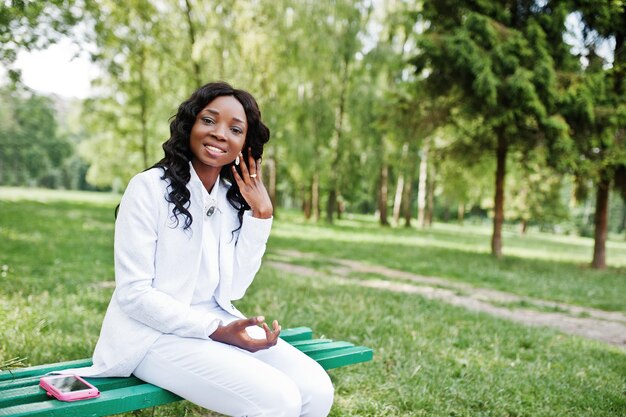 Ciérrese encima del retrato de la muchacha afroamericana negra elegante y hermosa que se sienta en banco con el teléfono móvil