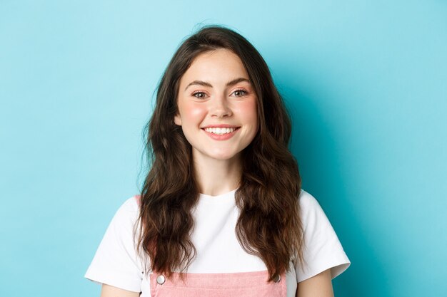 Ciérrese encima del retrato de la muchacha alegre del encanto con maquillaje lindo, dientes blancos sonrientes y que mira feliz a la cámara, de pie sobre fondo azul.