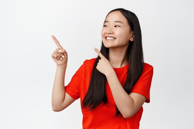 Ciérrese encima del retrato de la muchacha asiática hermosa con el pelo largo sano y la piel limpia que sonríe mirando y que señala en el fondo blanco del anuncio de la esquina superior izquierda