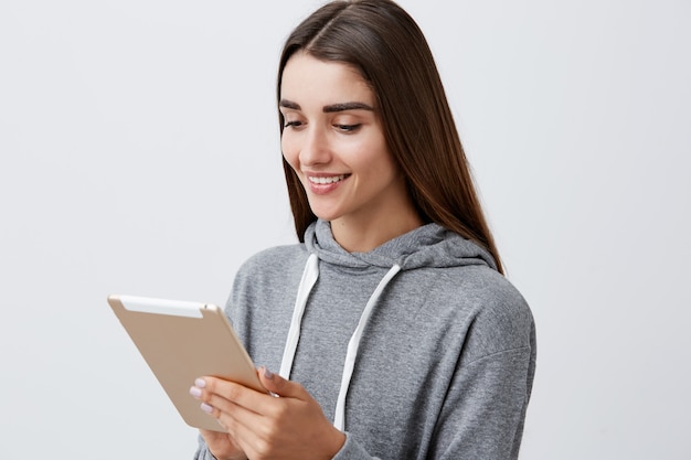 Foto gratuita ciérrese encima del retrato de la muchacha atractiva alegre del estudiante caucásico con el pelo largo oscuro n sudadera con capucha gris casual que sostiene la tableta digital en manos, mirando videos divertidos en línea con la cara feliz y relajada