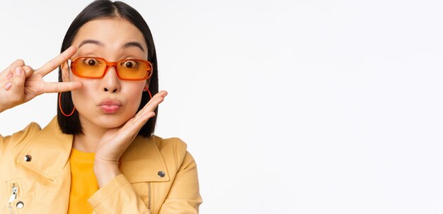 Ciérrese encima del retrato de la muchacha elegante de la mujer asiática joven en gafas de sol que muestran el signo de la paz y la sonrisa de pie sobre el fondo blanco del estudio