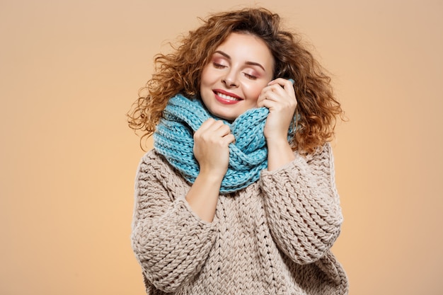 Foto gratuita ciérrese encima del retrato de la muchacha rizada morena hermosa soñadora alegre sonriente en suéter hecho punto y calentador gris sobre la pared beige