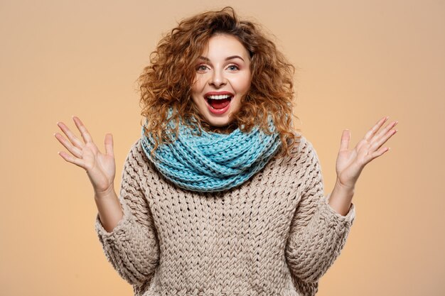 Ciérrese encima del retrato de la muchacha rizada morena hermosa sonriente alegre alegre en suéter hecho punto y el calentador gris sobre la pared beige