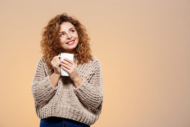 Ciérrese encima del retrato de la muchacha rizada morena hermosa sonriente alegre en suéter hecho punto que sostiene la taza sobre la pared beige