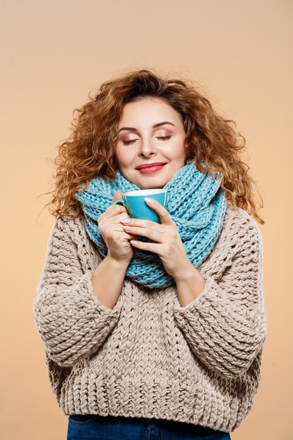 Ciérrese encima del retrato de la muchacha rizada morena sonriente hermosa alegre en suéter hecho punto y el calentador gris que sostiene la taza sobre la pared beige