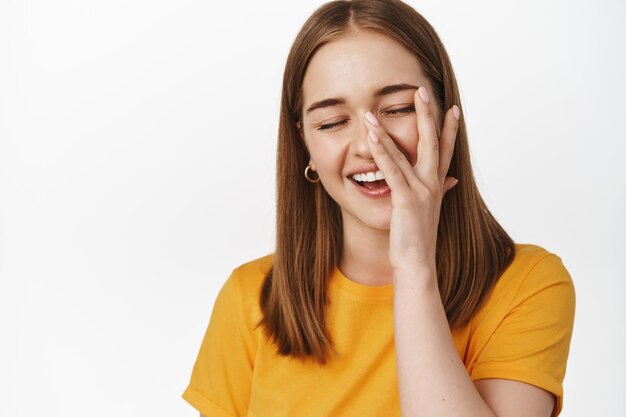 Ciérrese encima del retrato de la muchacha sincera feliz, sonriendo y riendo, tocando la piel facial con la mano, concepto de publicidad del cuidado de la piel y del tratamiento cosmético, fondo blanco