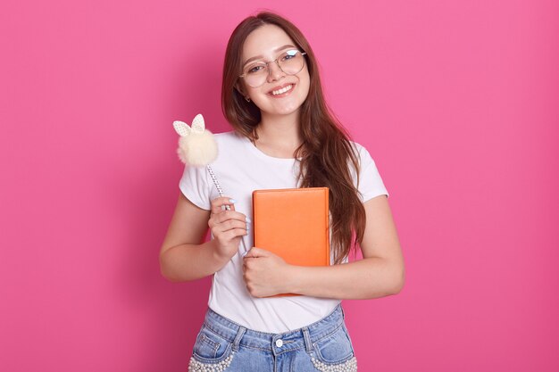 Ciérrese encima del retrato de la mujer atractiva joven que sostiene su cuaderno aislado sobre rosado