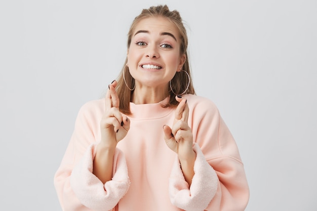 Ciérrese encima del retrato de la mujer bastante supersticiosa joven en sudadera rosada que mira la cámara que cruza sus dedos para la buena suerte. Chica estudiante esperando que sus sueños se hagan realidad. Expresiones faciales humanas