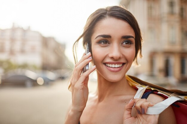 Ciérrese encima del retrato de la mujer caucásica joven alegre con el pelo oscuro que sonríe con los dientes, mirando a un lado con la expresión feliz y tranquila de la cara, hablando por teléfono con un amigo, sosteniendo bolsas de compras en