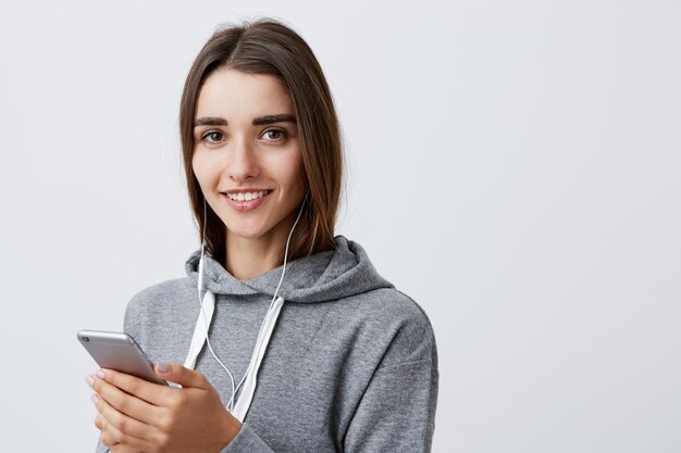 Ciérrese encima del retrato de la mujer caucásica morena feliz hermosa en sudadera con capucha gris casual que sonríe, sosteniendo el teléfono celular en manos, escuchando música en auriculares