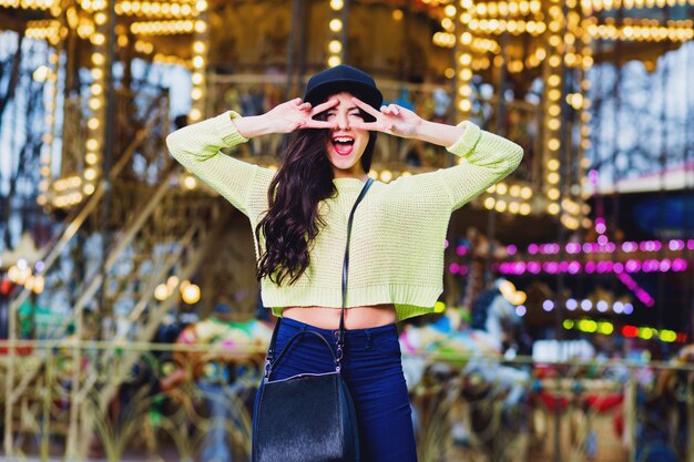 Ciérrese encima del retrato de la mujer elegante elegante atractiva diviértase y sonría en atracciones. Vistiendo moda hipster botín sombrero negro y suéter de neón.