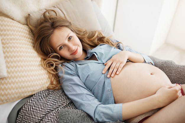 Ciérrese encima del retrato de la mujer embarazada hermosa joven alegre en la ropa casera con el pelo rizado claro que pone en cama con la expresión feliz y relajada, disfrutando de momentos de la maternidad.
