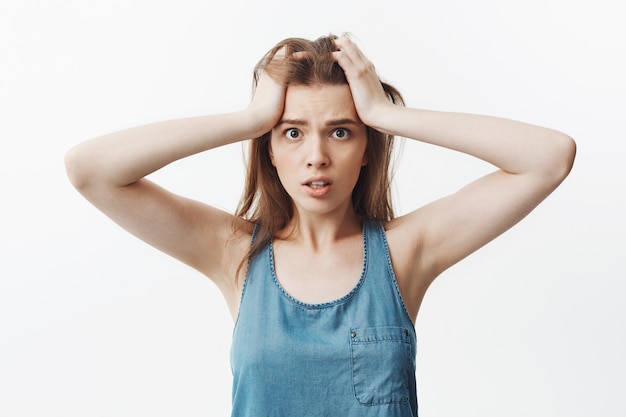 Ciérrese encima del retrato de la mujer europea morena joven hermosa en camiseta azul casual que aprieta la frente con las manos, con la expresión de la cara confundida y perdida.