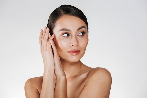 Ciérrese encima del retrato de la mujer hermosa con maquillaje natural que mira a un lado y que lleva a cabo las manos cerca de su cara sana, aislada sobre blanco