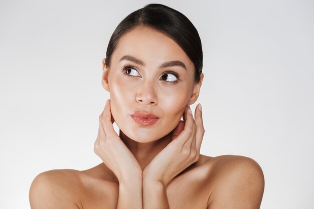 Ciérrese encima del retrato de la mujer hermosa con maquillaje natural que mira a un lado y que lleva a cabo las manos cerca de su cara sana, aislada sobre blanco