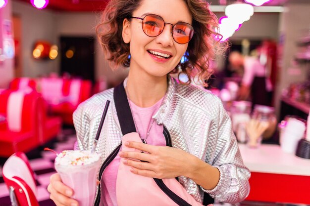 Ciérrese encima del retrato de la mujer hermosa sonriente con estilo en el café retro de los años 50 que bebe el cóctel del batido de leche en la chaqueta de plata de la camiseta rosa que lleva las gafas de sol rosadas que se divierten en humor alegre