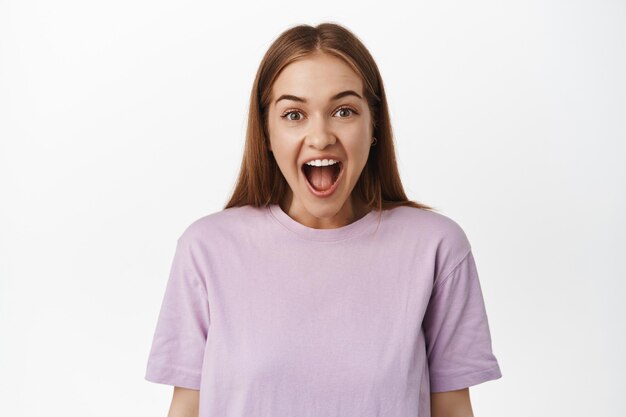 Ciérrese encima del retrato de la mujer impresionada y emocionada, deje caer la mandíbula por la emoción y el asombro, sonriendo ante la oferta de promoción impresionante, mirando al frente, de pie en una camiseta contra la pared blanca.