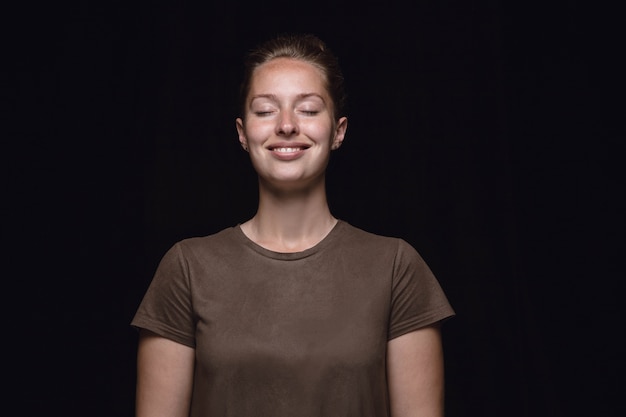 Ciérrese encima del retrato de la mujer joven aislada en el espacio negro. Photoshot de emociones reales de modelo femenino con los ojos cerrados. Pensando y sonriendo