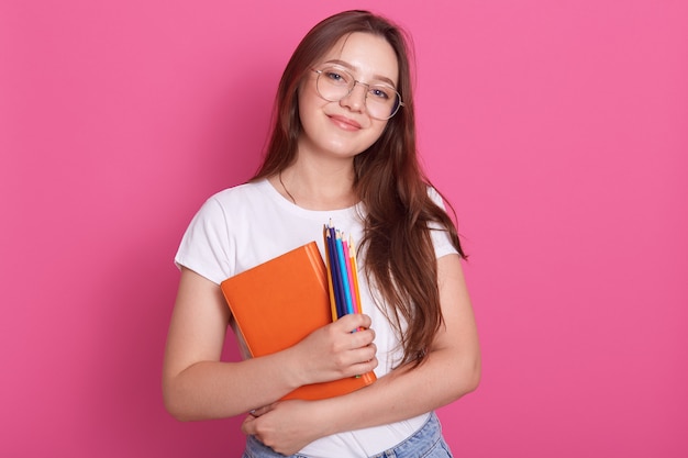 Ciérrese encima del retrato de la mujer joven linda que sostiene el libro de texto y los lápices de colores, posando en el estudio aislado sobre rosa