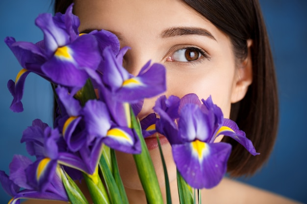 Ciérrese encima del retrato de la mujer joven tierna detrás de los iris violetas sobre la pared azul