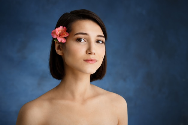 Ciérrese encima del retrato de la mujer joven tierna con la flor rosada en pelo sobre la pared azul