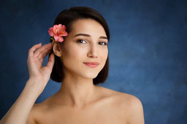 Ciérrese encima del retrato de la mujer joven tierna con la flor rosada en pelo sobre la pared azul