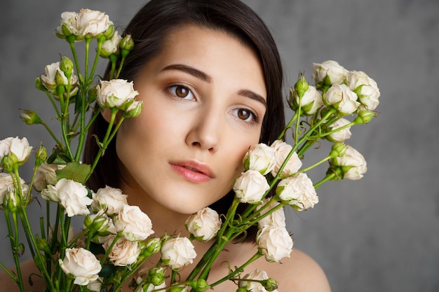 Ciérrese encima del retrato de la mujer joven tierna con las flores sobre la pared gris