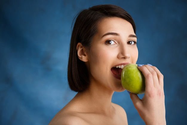 Ciérrese encima del retrato de la mujer joven tierna que muerde la manzana sobre la pared azul