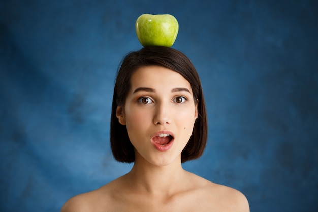 Foto gratuita ciérrese encima del retrato de la mujer joven tierna que sostiene la manzana en su cabeza sobre la pared azul