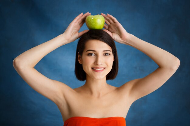 Ciérrese encima del retrato de la mujer joven tierna que sostiene la manzana en su cabeza sobre la pared azul
