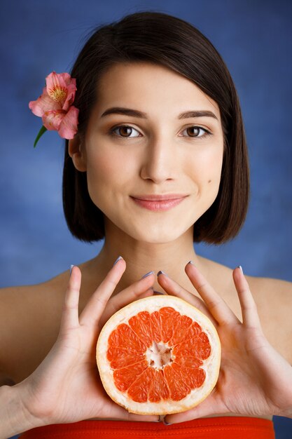 Ciérrese encima del retrato de la mujer joven tierna que sostiene la naranja cortada sobre la pared azul