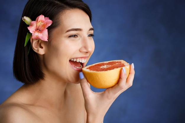 Foto gratuita ciérrese encima del retrato de la mujer joven tierna que sostiene la naranja cortada sobre la pared azul