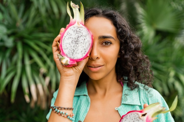 Foto gratuita ciérrese encima del retrato de la mujer morena encantadora sonriente con los pelos ondulados que sostienen las frutas tropicales