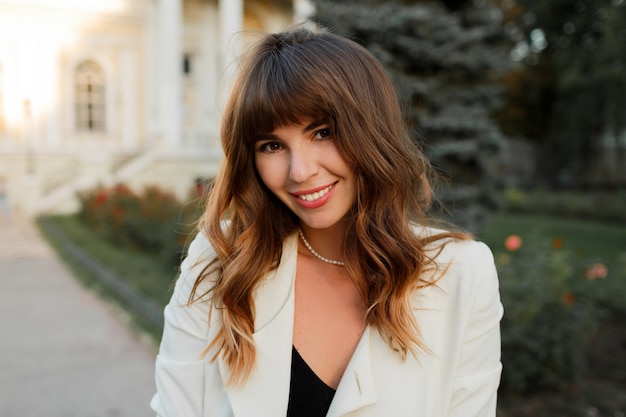 Ciérrese encima del retrato de la mujer de negocios exitosa atractiva que presenta al aire libre. Cabello ondulado, elegante chaqueta blanca.