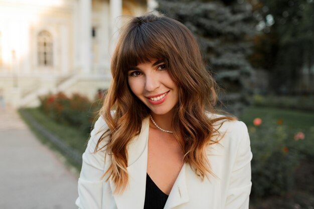 Ciérrese encima del retrato de la mujer de negocios exitosa atractiva que presenta al aire libre. Cabello ondulado, elegante chaqueta blanca.