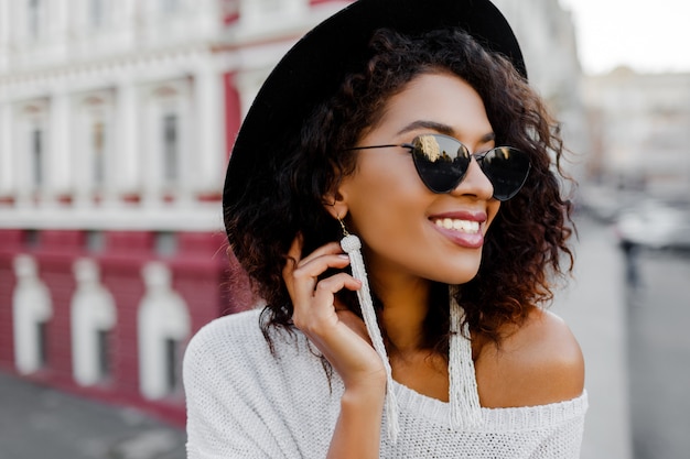 Ciérrese encima del retrato de la mujer negra de moda con la presentación elegante de los pelos del Afro al aire libre. Fondo urbano Lleva gafas de sol negras, sombrero y aretes blancos. Accesorios de moda. Sonrisa perfecta.