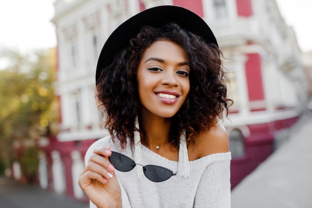 Ciérrese encima del retrato de la mujer negra de moda con la presentación elegante de los pelos del Afro al aire libre. Fondo urbano Lleva gafas de sol negras, sombrero y aretes blancos. Accesorios de moda. Sonrisa perfecta.