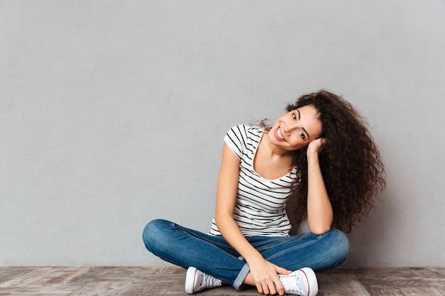 Ciérrese encima del retrato de la mujer rizada en ropa casual que se sienta en la postura del loto en el piso apoyando su cabeza con la mano feliz y sincera sobre la pared gris