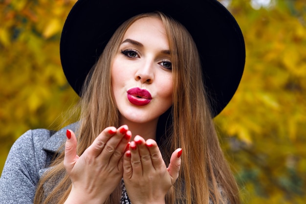 Ciérrese encima del retrato de la mujer rubia bonita elegante que presenta en el día frío del otoño en el parque de la ciudad, vistiendo sombrero negro elegante, pelo largo, maquillaje brillante. Enviando beso al aire