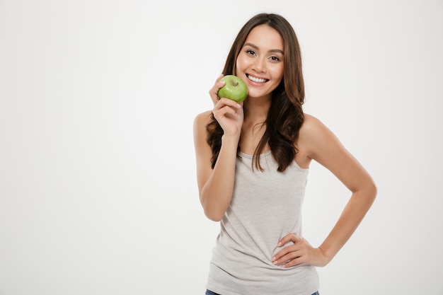 Ciérrese encima del retrato de la mujer sonriente con el pelo castaño largo que mira en cámara con la manzana verde en la mano, aislada sobre blanco