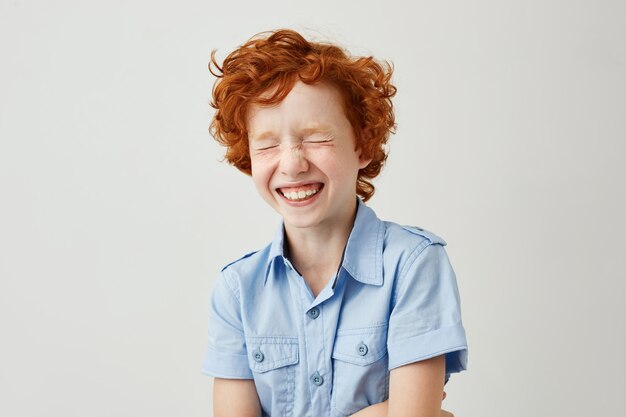 Ciérrese encima del retrato del niño alegre con el pelo rizado del jengibre y las pecas que ríen con los ojos cerrados