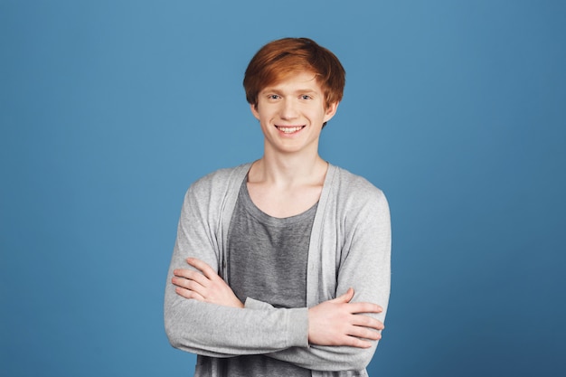 Ciérrese encima del retrato o estudiante masculino pelirrojo alegre atractivo joven en traje gris casual que sonríe con los dientes, cruzando las manos, con la expresión confiada y feliz.