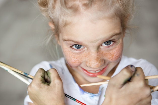 Ciérrese encima del retrato de la pequeña niña rubia adorable en la camiseta blanca que sostiene cepillos, divirtiéndose, disfrutando de dibujar con expresión feliz