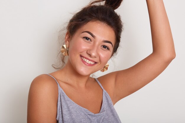 Ciérrese encima del retrato de la señora linda y sonriente con el pelo ondulado oscuro, chica joven con la cola de caballo mujer adorable que lleva la camiseta casual gris aislada sobre la pared blanca del estudio.