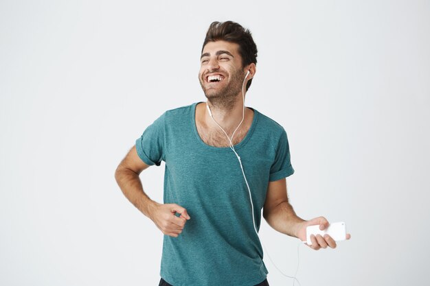 Ciérrese encima del retrato del tipo caucásico hermoso divertido en camiseta azul, escuchando música en auriculares, engañando y bailando con expresiones felices y ojos cerrados.