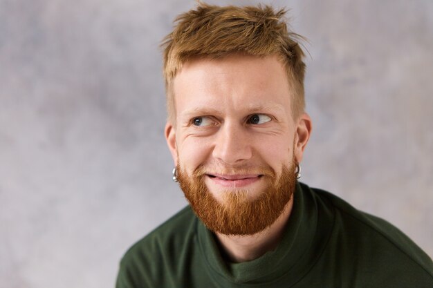 Ciérrese encima del retrato del varón caucásico joven hermoso con el peinado elegante y la barba recortada que presenta aislado que mira lejos con la sonrisa misteriosa, sintiéndose muy halagado. Expresiones humanas positivas