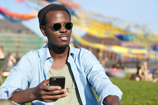 Ciérrese encima del retrato del varón de piel oscura de moda en gafas y camisa de moda, sosteniendo el teléfono celular en la mano, mirando en la distancia mientras se relaja en la hierba verde al aire libre. Personas, estilo de vida, tecnología.