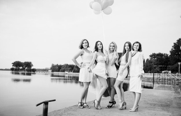 Cinco chicas con globos en la mano vestidos de blanco en despedida de soltera contra el muelle en el lago