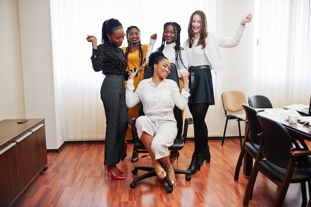 Cinco mujeres de negocios multirraciales de pie en la oficina y ruedan a la mujer en la silla Diverso grupo de empleadas en ropa formal divirtiéndose